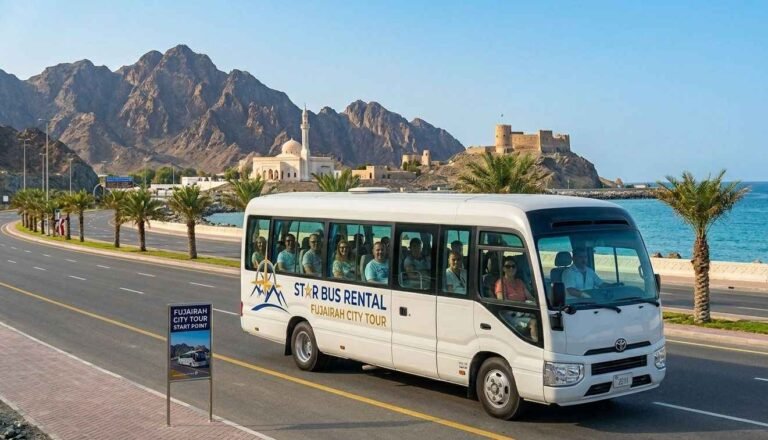 White Toyota Coaster minibus with passengers and driver on a Fujairah city tour by Star Bus Rental, driving along the coastal road with the Hajar mountains and Fujairah Fort in the background